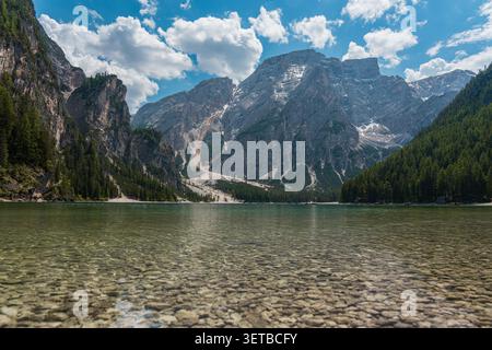 Kristallklares, seichtes Wasser des Pragser Sees mit einem Kieselsteinbett mit hoch aufragenden felsigen Berggipfeln und grünen Kiefernwäldern unter sonnigem Himmel, sce Stockfoto