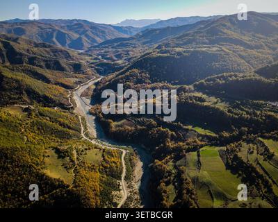 Dieser Blick aus der Vogelperspektive fängt einen sich windenden Fluss und eine unbefestigte Straße ein, die sich durch ein riesiges, bewaldetes Bergtal schlängelt. Die Landschaft zeigt Herbstlaub mit Stockfoto