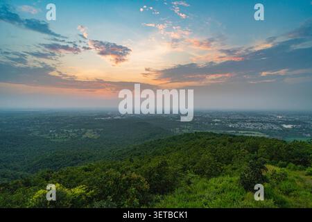 Ein atemberaubender Blick auf einen lebendigen Sonnenuntergang, der warme Farbtöne über einem dichten grünen Wald und einer weitläufigen Stadt in Rajasthan, Indien, ausstrahlt. Stockfoto