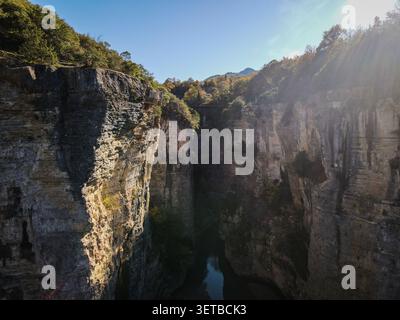 Ein dramatischer Blick aus der Luft fängt eine tiefe Schlucht mit hoch aufragenden Kalksteinklippen auf beiden Seiten ein, die teilweise von hellem Sonnenlicht beleuchtet werden. Ein schmaler Fluss fl Stockfoto