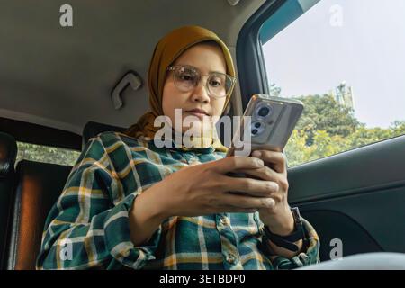 Eine asiatische muslimische Frau, die einen Hijab und eine Brille trägt und eine Nachricht auf ihrem Handy schreibt, während sie mit dem Auto reist Stockfoto