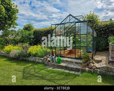Glasgewächshaus in einem üppigen Garten im Hinterhof mit Blumenrändern und sonniger Sommerlandschaft Stockfoto
