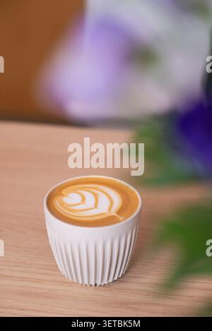 Ein frisch gebrühter heißer Cappuccino mit detaillierter Latte-Kunst, serviert in einer weißen gerippten Keramikschale auf einem natürlichen Holztisch. Stockfoto