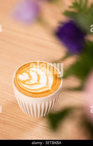 Ein frisch gebrühter heißer Cappuccino mit detaillierter Latte-Kunst, serviert in einer weißen gerippten Keramikschale auf einem natürlichen Holztisch. Stockfoto