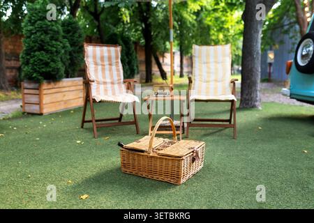 Gemütlicher Picknickbereich im Freien mit zwei Klappstühlen aus Holz, gestreiften Kissen, einem kleinen Tisch mit einem Vintage-Radio und einem Korb auf einem grünen Rasen. Stockfoto