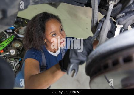 Afroamerikanische Mechanikerin inspiziert mechanische Teile eines Fahrzeugs, während sie es in einer Autowerkstatt repariert. Werkzeuge befinden sich in der Nähe. Stockfoto