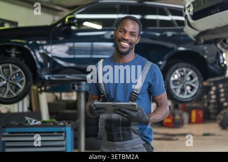 Porträt eines lächelnden Automechanikers oder Fahrzeugtechnikers in einem Arbeitsanzug und mit Sicherheitshandschuhen, mit einem Tablet in einer modernen Werkstatt. Stockfoto