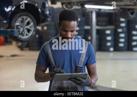 Afroamerikanischer Mechaniker mit einem Tablet zur Diagnose von Pannen, Überprüfung der Wartung und Bestellung von Teilen neben angehobenen Autos, Porträt. Stockfoto