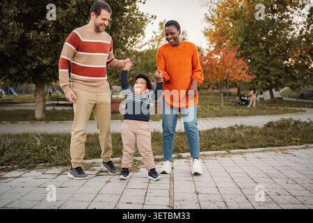 Interrassische Eltern gehen mit glücklichem Kleinkind-Sohn, der die Hände hält Stockfoto