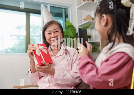 Asiatische Großmutter hält eine Geschenkbox in der Hand, während die Enkelin während der Geburtstagsfeier zu Hause Fotos mit dem Smartphone macht Stockfoto