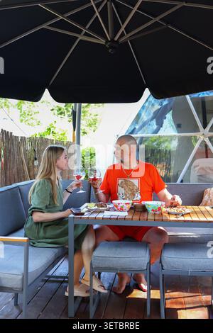 Glückliches Paar, das während eines entspannten Urlaubs auf einer gemütlichen Terrasse in der Nähe einer geodätischen Kuppel mit Rotwein tobt. Stockfoto