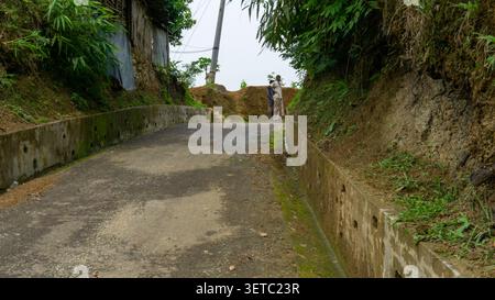 Betonweg mit Entwässerungslöchern, die an einfachen Bambushäusern vorbeiführen. Männer, die in der Ferne stehen. Hügeliges Gelände mit dichter grüner Vegetation und Bäumen. Stockfoto