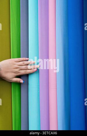 Eine weibliche Hand mit farbenfroher Maniküre, die mit leuchtendem vertikalem Stoff in Berührung kommt, rollt in einem Geschäft. Konzept: Auswahl haptischer Materialien und individuelles Modedesign Stockfoto