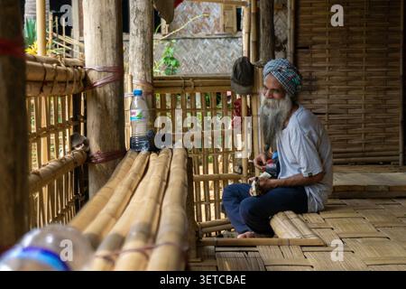 Seitenprofil-Aufnahme eines älteren Mannes mit weißem Bart und Turban, der in traditionellem Haus auf einem gewebten Bambusboden sitzt. Stockfoto