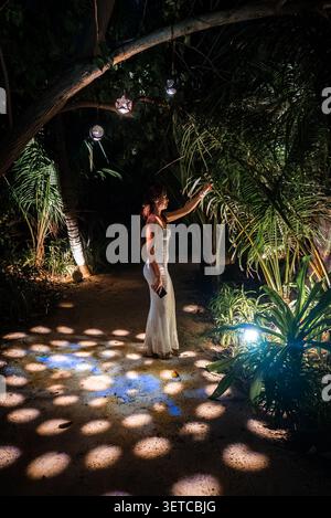 Frau in weißem Kleid geht nachts auf den Malediven durch Palmen gesäumte Gartenwege Stockfoto
