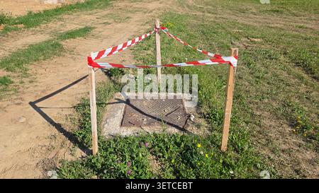 Ein Deckel aus Metall auf einem Betonuntergrund ist von einem temporären Sicherheitsband aus gestreiftem Band und Holzpfählen in einem grasbewachsenen Gelände umgeben Stockfoto