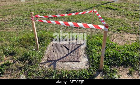 Ein kaputter Metalldeckel auf einem Betonsockel ist mit Sicherheitsstreifen und Holzpfählen auf einem grasbewachsenen Feld abgesichert Stockfoto