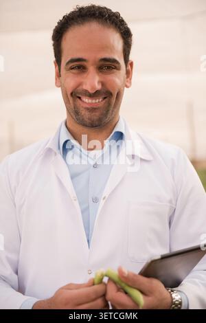 Zuversichtlicher nahöstlicher Ernährungsberater oder Ernährungsberater im weißen medizinischen Mantel, der warm lächelt, während er frischen grünen Apfel und Klemmbrett im Freien hält Stockfoto