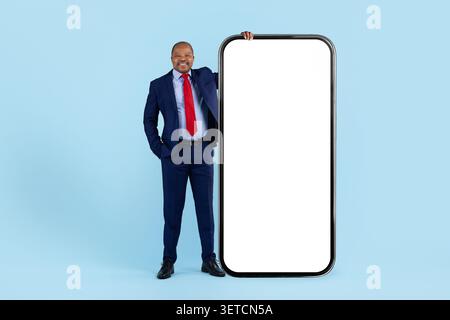 Lächelnder reifer schwarzer Geschäftsmann im Anzug, der sich gegen ein übergroßes leeres Smartphone-Modell lehnt Stockfoto