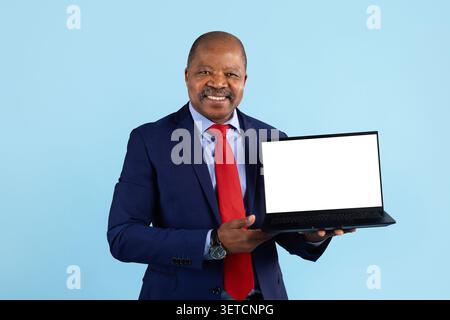 Lächelnder, reifer schwarzer Geschäftsmann im Anzug, der einen Laptop mit leerem Bildschirm hält Stockfoto