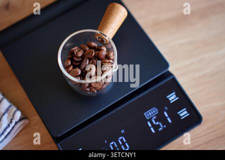 Geröstete Kaffeebohnen in einer Glaskugel auf einer schwarzen Digitalskala auf einem Holztisch. Konzept der Kaffeezubereitung, Messung von Bohnen und Tropfkaffee-Brauerei Stockfoto