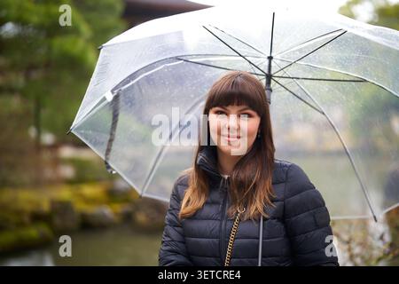 Lächelnde junge Frau mit klarem Schirm in einem regnerischen japanischen Garten in Kyoto Stockfoto