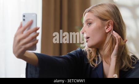 Fröhliche kaukasische Frau lächelt und fotografiert Selfie mit Handy-Grimassen im Schlafzimmer. Unbeschwertes Fotografieren von Frauen mit Smartphone Stockfoto
