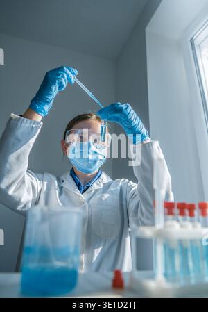 Eine Wissenschaftlerin mit Handschuhen, Maske und Schutzbrille arbeitet in einem hellen Labor mit blauen Chemikalien. Sie transferiert vorsichtig Flüssigkeit mit einer Pipette und demonstriert fortschrittliche Forschungstechniken Stockfoto