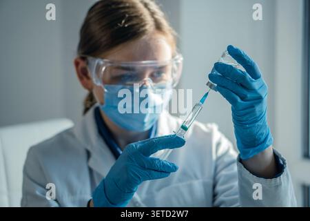 Ein Arzt in blauen Handschuhen und Laborkittel zieht Flüssigkeit aus einer Durchstechflasche in eine Spritze, wobei der Schwerpunkt auf Genauigkeit liegt. Das Bild zeigt moderne Gesundheits-, Impfungs- und Sicherheitsprotokolle Stockfoto