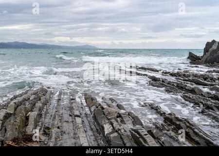 Wellen brechen unter bewölktem Himmel an der Küste von Zarautz, Spanien, gegen die geschichteten Felsformationen Stockfoto