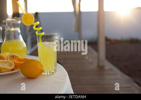 Glas gefüllt mit blassgelber Limonade und Eis, lockige Zitronenscheibe Strohgarnitur, ganze Zitrone und halbe Zitronen auf weißer Tischdecke, Krug Limonade hinein Stockfoto