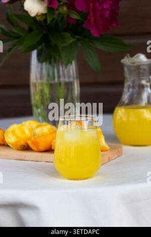 Hellgelbe Zitruslimonade in kurzes Glas mit Eis auf weißer Tischdecke gegossen, hölzerne Schneidebrett mit halbierten Zitronen und ganzen Zitronen hinein Stockfoto