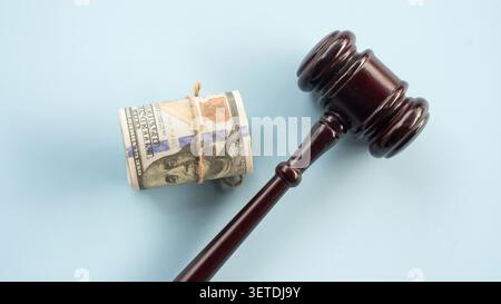 Hölzerner richterlicher Hamel, der neben einer Rolle von US-100-Dollar-Scheinen auf einem soliden, schlichten blauen Hintergrund für Rechtskosten und Korruption liegt Stockfoto