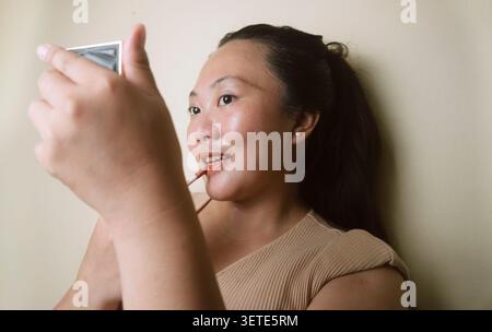 Junge Frau, die Lippenstift aufträgt, während sie drinnen in einen Handspiegel blickt. Stockfoto