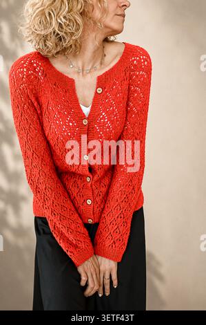 Fröhliche blonde Frau im hellen roten Strickpullover, der barfuß auf Holzboden sitzt. Glückliche Blondine. Lockiges Haar. Strickjacke in Orange. Stockfoto