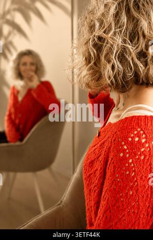 Fröhliche blonde Frau im hellen roten Strickpullover, der barfuß auf Holzboden sitzt. Glückliche Blondine. Lockiges Haar. Strickjacke in Orange. Stockfoto
