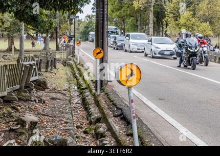 Wildwarnschilder neben dem Verkehr auf einer Straße durch den Nara-Park, wo freilebende Sikawildschweine in Nara, Japan, die Straße überqueren. Stockfoto