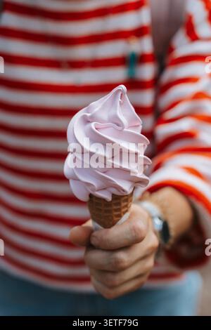 Frau mit einem Wirbel rosafarbenem Eis in einer Waffelschale, während die Person ein leuchtendes, rot-weiß gestreiftes Langarmshirt im verschwommenen Hintergrund trägt, das eine verspielte und stilvolle Sommeratmosphäre weckt. Stockfoto