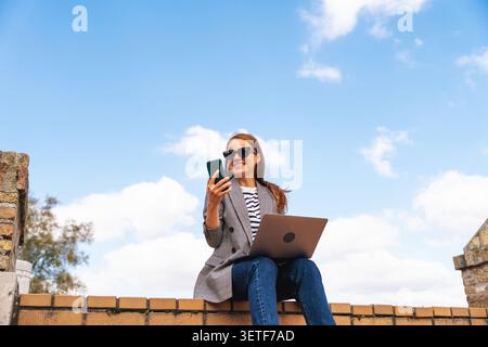 Frau, die Smartphone und Laptop benutzt, während sie ferngesteuert auf urbanen Stufen arbeitet Stockfoto