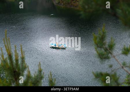 Ein kleines Ruderboot bringt zwei Personen über das gewellte Seewasser, mit dunkler Waldküste im Hintergrund und weichen Kiefernzweigen, die die Szene umrahmen Stockfoto