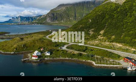 Aus der Vogelperspektive auf die gewundene Bergstraße durch die üppige grüne Lofoten-Landschaft mit traditionellen weißen und roten Häusern, Fischerbooten und dramatischen Stockfoto