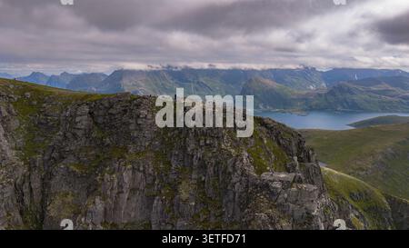 Dramatische Felsenklippen mit Wanderern, die die atemberaubende Landschaft des Fjordes der Lofoten Islands überblicken. Alpengipfel und blaues Wasser schaffen eine Stockfoto