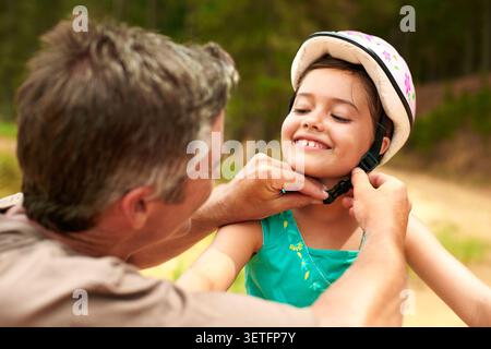 Lächeln, Vater und helfendes Kind mit Helm, Sicherheit und Anleitung für Fahrradfahren. Glücklich, Mann und Tochter in der Natur mit Schutzausrüstung, Radfahren Stockfoto