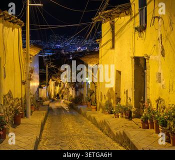 Eine steile, enge, gepflasterte Straße in Cusco, Peru, am späten Abend mit zwei Personen zu Fuß und mit den Lichtern der Stadt im Hintergrund. Stockfoto