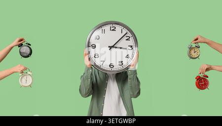 Person, die eine große Uhr vor dem Gesicht hält, mit mehreren Weckern, die von den Händen auf grünem Hintergrund gehalten werden Stockfoto