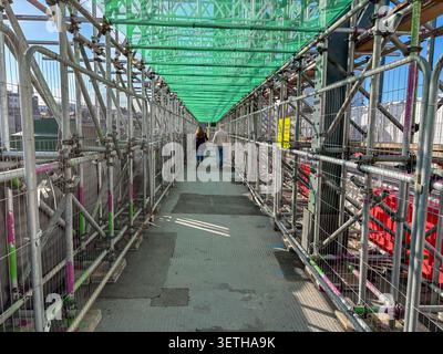 Fußgänger, die durch einen schmalen überdachten Gehweg aus Metallgerüsten und grünem Schutznetz auf einer Stadtstraße laufen. Stockfoto