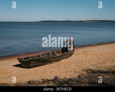 Verwittertes Holzruderboot liegt an einem sandigen Ufer.nCalm blaues Wasser erstreckt sich in Richtung entfernter Inseln.NA der Mann sitzt entspannt im Boot und trägt einen dunklen jac Stockfoto