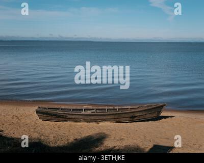 Ein altes Holzboot ruht an einem Sandstrand. Ruhiges blaues Wasser reicht bis zum Horizont. Sanfte Wellen umrunden das Ufer. Klarer Himmel mit weichen Wolken darüber. Sere Stockfoto