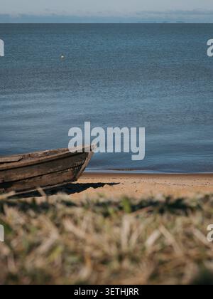 Verwittertes Holzboot ruht auf Sandstrand. Ruhiges blaues Wasser zieht sich in Richtung entfernter Hügel. Sonnenlicht leuchtet sanft auf die Wasseroberfläche. Getrocknetes Gras f Stockfoto