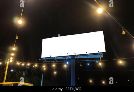 Ein großes leeres weißes Reklametafelmodell steht bei Nacht vor einem dunklen Himmel, umgeben von leuchtenden dekorativen Lichtern. Diese Outdoor-adve Stockfoto